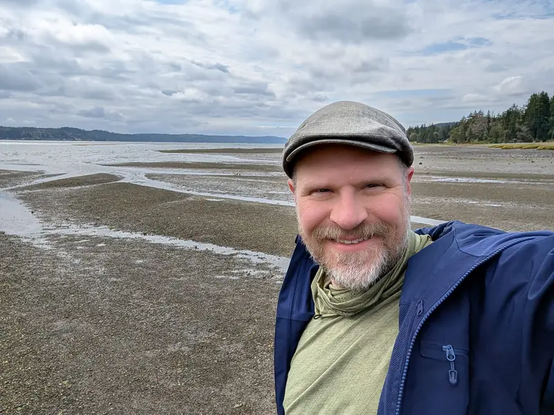 Smiling bearded man in a flat cap and navy jacket on a Puget Sound beach at low tide