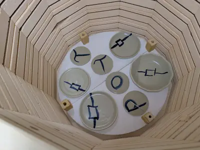 Eight pale green plates and dishes with blue brushed lantern motifs and letters, viewed from above inside a kiln